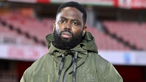 Ghetts wearing a green jacket looking serious and thoughtful.  An empty Emirates stadium in the  background. He has a short beard