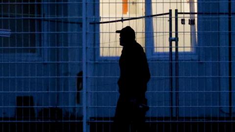 A man in a baseball cap walks past a window at night. He's silhouetted against the building and the soft white light coming from the window.
