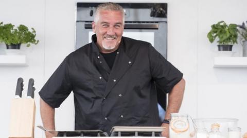 Paul Hollywood has short grey hair and is wearing a black t-shirt and a black chef's coat. He is stood in a kitchen has his hands on a work surface. A set of knifes and glass containers are in the foregrounds and an oven and two potted plants are in the background.