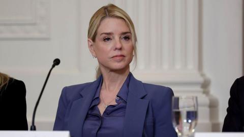 Former US Attorney General Pam Bondi sits at a table behind a tabletop microphone as she listens to US President Donald Trump speak during a lunch with the Trump Kennedy Center Board Members on 16 March 2026.