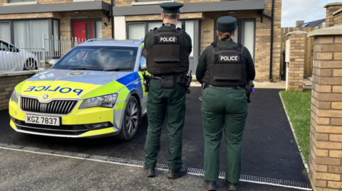 Shows a police car in yellow and blue with two police officers - a man and woman - standing with their backs to the camera 