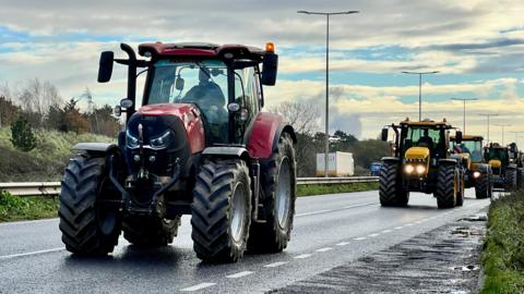 A convoy of farm vehicles moving down a dual carriageway.  There is a red tractor at the front with three yellow-coloured vehicles behind it.