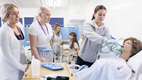 Five women in a hospital-like learning environment. One, who has brown hair and is wearing a jumper is putting a blue tube into a doll's mouth. Two blonde women are watching. Two of the women are in the background.