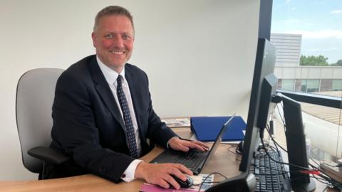 A picture of new Conservative Buckinghamshire council leader Steven Broadbent. He is sat down in front of his laptop and desk, and is wearing a black suit jacket with a white shirt and black tie with red spots.