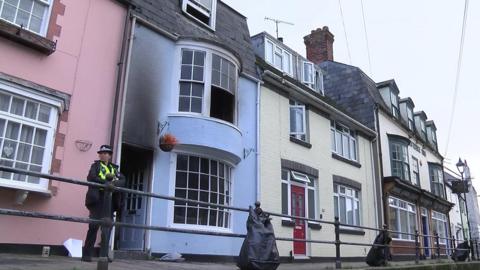 A female police officer standing outside a blue terraced property with the front door open and signs of fire damage above it.