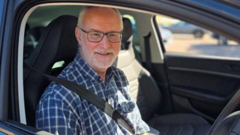 Fran Whymark, a man in a blue chequered shirt who is sitting inside a car in the driving seat and is wearing a seat belt. 