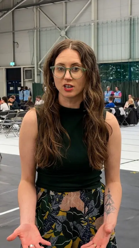 Woman with long brunette hair wearing glasses presenting to camera