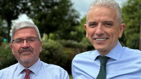 Nick Mannion, a man with grey hair and glasses and wearing a blue shirt and red tie, is standing next to Michael Gorman, a man with grey hair wearing a blue shirt and green tie. They are outdoors - with trees visible in the background.