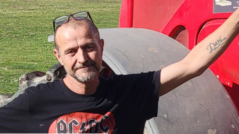 David Evans standing next to an agricultural vehicle in a field. He has short hair and a goatee beard and is wearing a red and black AC DC t-shirt 