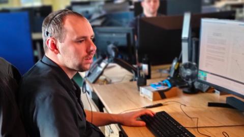 A man sitting at a desk working as an emergency call handler in a control room for Norfolk Fire and Rescue Service. He is wearing a black short sleeved shirt and is looking towards a computer screen.