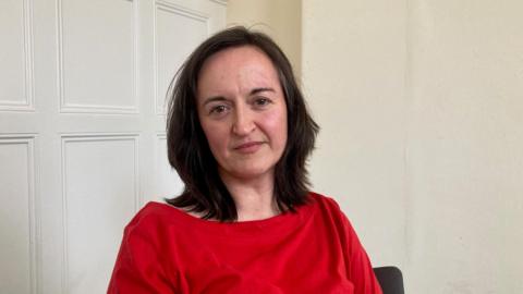 A woman with long, dark hair wearing a red top and sitting in a room with a white panelled wall behind her