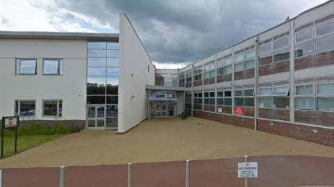 A Google Streetview image of a school on a day with grey skies. It is a rendered building with glass windows along the top and bottom levels. 