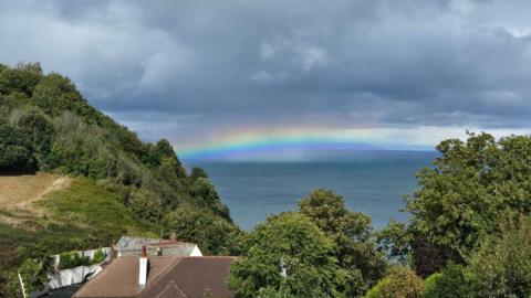 Rainbow on the horizon above the sea with rain clouds above, house roof and topiary in the foreground