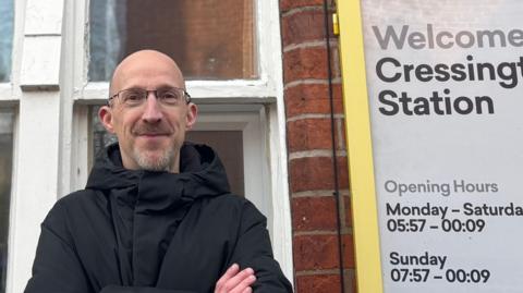 Councillor Richard Clein with a grey beard wearing glasses and a black coat stands at Cressington railway station with a yellow sign showing opening hours to his right.