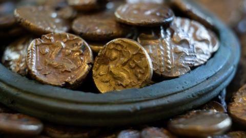 A close-up of a pile of 933 Iron Age gold coins. It shows the rim of a vessel, surrounded by coins with more gold coins heaped within.
