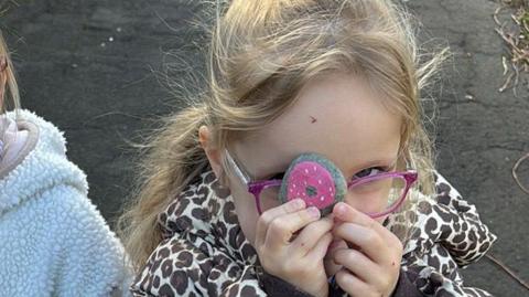 A little girl is holding up a pink painted stone. She has long blonde hair and pink spectacles.