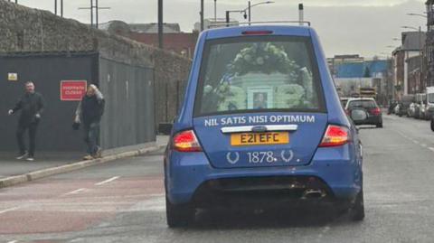 The back of a blue funeral car with a white coffin inside covered in flowers. The name "Rob" is spelled in white flowers. Nil Satis Nisi Optimum (the Everton Motto - Latin for Northing But The Best Is Good Enough) is written in white letters on the back of the car.