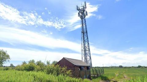 A telecommunications mast stands in a field next to a small fenced off building.