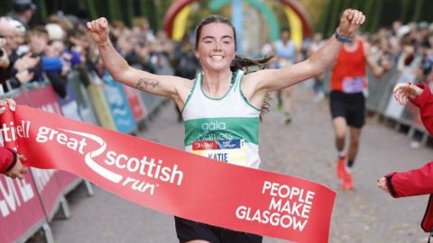 a runner wearing a number bib that reads 'Katie' and a white and green vest that says 'Gala Harriers', crosses the finish line of the Great Scottish Run with arms raised. The finish tape reads "People Make Glasgow".
