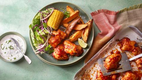 A green plate filled with potato wedges, corn on the cob and glazed chicken chunks with salad on a green surface