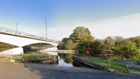 Road with double yellow lines and slipway with river behind and road bridge over the water