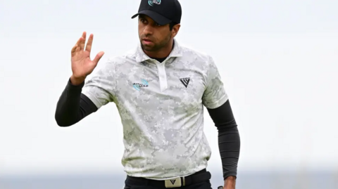 Rai is wearing golf gear and raising his right hand as a wave possibly to spectators. He has a white top on over a long sleeve black top and a white cap.