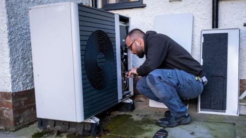 A man crouches down to carry out work on a heat pump, which is fixed to the ground outside a property. He is wearing a black hooded top, denim combat-style trousers and black trainers. He has cropped dark hair, a beard and is wearing glasses.