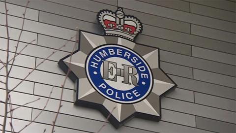 The Humberside Police badge on the wall of a police station. It has the words Humberside Police in white letters in a circle with a blue background and the initials ER in the middle. A crown sits on top of the badge