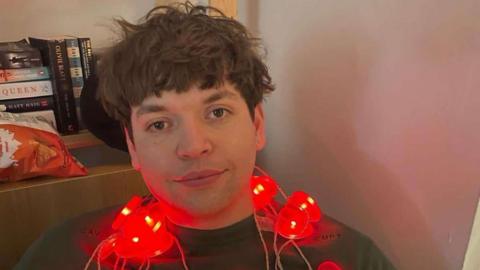 Luis Piovesana wearing a dark grey jumper and sitting down in a corner, leaning up against a wooden bookcase. He has a string of red, love-heart shaped fairy lights around his neck. He has a dark brown, shaggy haircut and brown eyes, and is softly smiling at the camera.