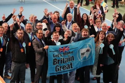 A group of Reform councillors celebrate the election result in Sunderland. There is a group of men and women with their arms in the air. Those at the front are holding a banner which has a Reform UK logo and the words Let's Make Britain Great