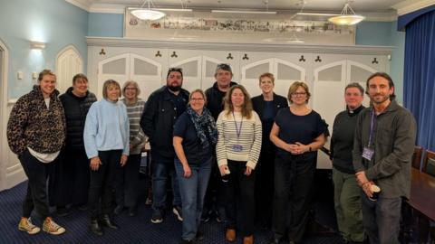 A group picture of a dozen or so individuals standing in a room with a blue carpet and blue and white walls.