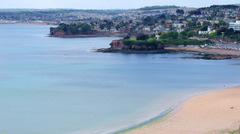 The picture shows a wide, calm bay with blue water stretching out towards a sandy beach in the foreground. The beach curves gently along the shoreline, with only a few tiny figures walking near the water’s edge. The sea looks still and clear, with light ripples close to the coast. Further out, a rocky headland covered in green trees juts into the water.