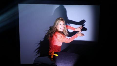 Presenter Naomi Wilkinson in a red jumper. She is holding her hands side by side creating a shadow against a blue background.