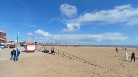 A Google street view of Skegness Promenade looking out at the beach and sea with the pier in the distance. You can see several people walking or sitting and an ice cream van in the middle.