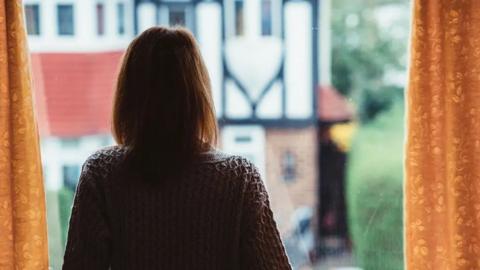 Generic shot of the back of a woman with brown hair cur in a bib, wearing a dark-coloured woolen cardigan, looking out of a window towards a house with white render across much of the front with brown edging. The window of the woman's house has yellow curtains. A hedge is visible to the right.