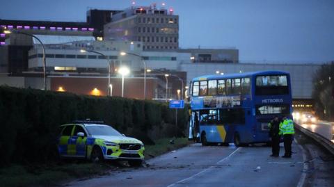 A blue double-decker bus is across a carriageway on an airport road and the door is hanging off. A police car is nearby on a verge with the back of the vehicle in a hedge and damage to the front of the marked vehicle. Two police officers with hi-vis jackets are standing near the bus. One is looking at a notebook and the other is talking on his phone.