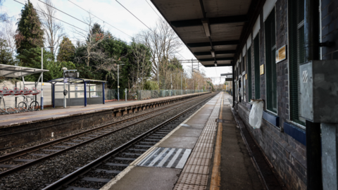 An image of a train station platform. It is surrounded by trees. It has two railway lines, two platforms and a brick waiting room with a small canopy to shelter the passengers. There is also a bike rack and a perspex shelter on the opposite platform.