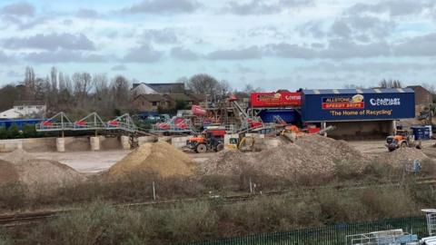 A stone crushing plant with a lot of heavy machinery on a churned up site. Houses can be seen behind it and in front of the site, where a lot of crushed up stone has been placed in piles, there is a railway line. The sky is blue with little grey clouds scattered across it.