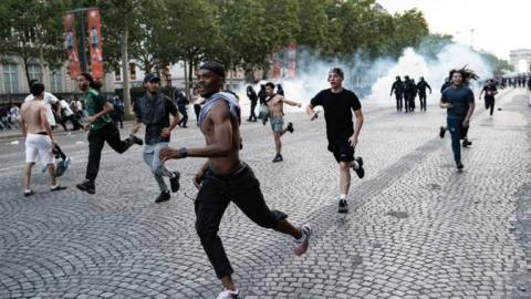 Young men run away from police on a cobbled street. Smoke from tear gas can be seen in the backgound