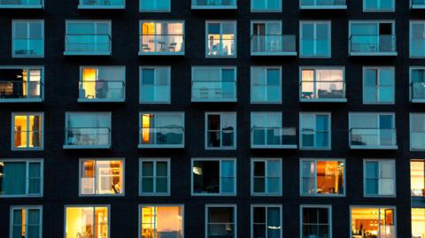 Stock image of a modern building facade in London at night - it shows residential flats where there are five rows of windows in the shot, some with curtains closed and lights off and some with lights on. 