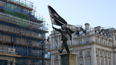 A statue is pictured showing a man holding a flag walking off a plinth. People are surrounding it, taking pictures of it. The statue is placed against the backdrop of historic buildings.