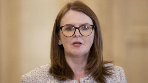 Caoimhe Archibald in glasses at a broadcast microphone. She is wearing glasses and textured blazer, and is in the middle of speaking