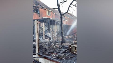 Embers and burnt struts belonging to a porch-type structure at the back of a house. There is a blackened tree and smoke is emanating from the ground. Broken glass and debris lie on the ground along with burnt fence panels. 
