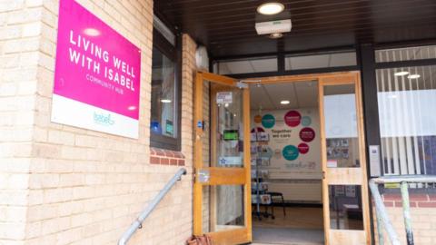 A brick building with an open door. On the wall of the building is a magenta and white sign, with "Living well with Isabel" on it in capital letters. Inside the open door, on a back wall, is a sign that says "Together we care".