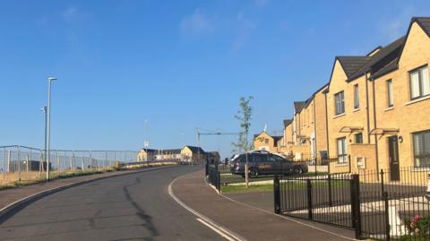A line of houses which are light coloured. There is a metal fence on the other side of the road and a shrubby area.