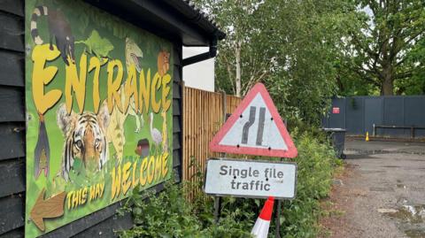 Shepreth Wildlife Park is on the left and has a sign on the side of the building which says ENTRANCE and an arrow pointing left. There is a cone and single file traffic sign outside of the building.