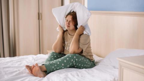 A woman sat on her bed in her pyjamas with a pillow wrapped around her ears. She is grimacing, as if being disturbed by noise.