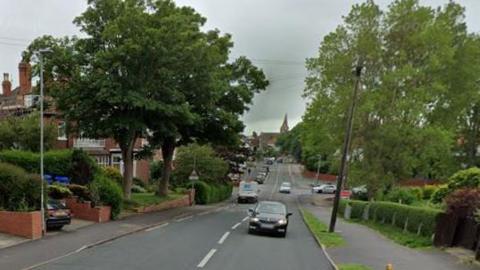 Tree-lined road with houses, lamp-posts, signs and cars in view