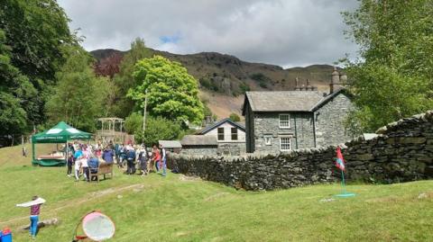 Langdale CofE Primary School in the Lake District, Cumbria. The school building is built with picturesque blue slate. It is in a green environment and has a dry stone wall surrounding the premises. Its grassy grounds are filled with parents and children on what looks like a sports day.