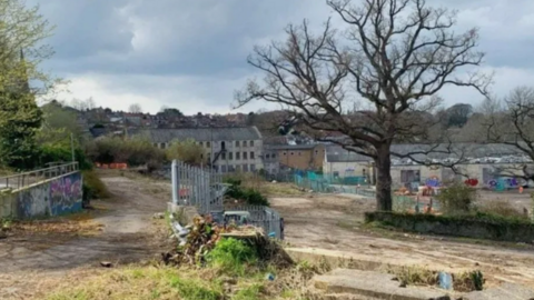 Saxonvale site in the middle of Frome looking derelict. Tree in the foreground and old empty factory buildings in the background There are bits of greenery among the paths. There are a couple of sets of railings, one of them above a wall of graffiti.